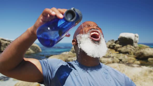 Senior african american man exercising pouring water on face on rocks by the sea