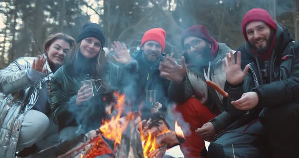 Happy Young Friends Sitting By Campfire in Winter Forest and Waving Hands at Camera