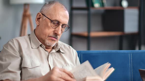 Elderly Male Reading Interesting Paper Book Turning Pages Smiling Enjoy Retirement at Home Closeup