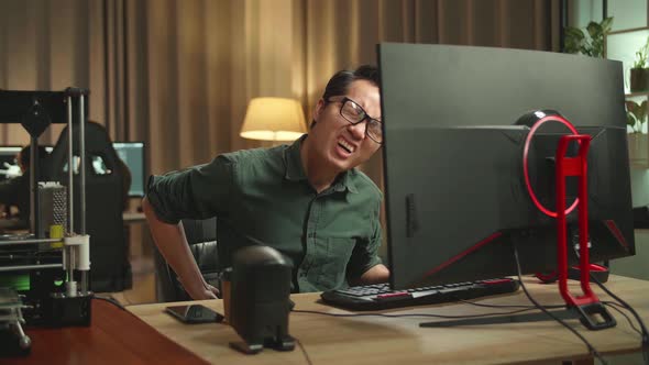 Asian Man Paining His Back While Works On Personal Computer And 3D Printer In Home Office