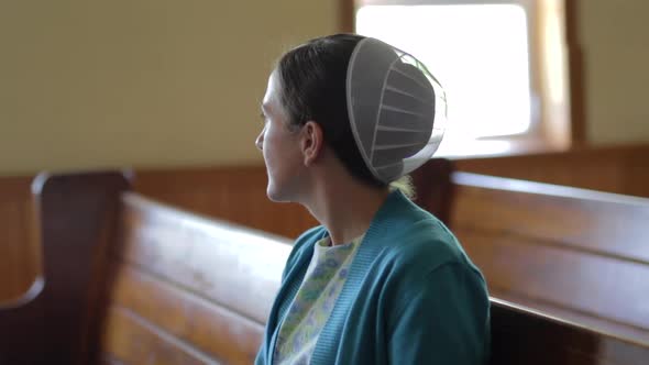 A portrait of a young Mennonite woman looking out a church window.