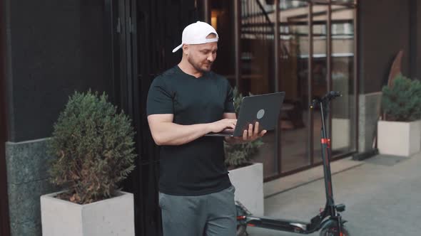 Adult man well dressed with electric scooter working with his laptop in the street in the city. A yo