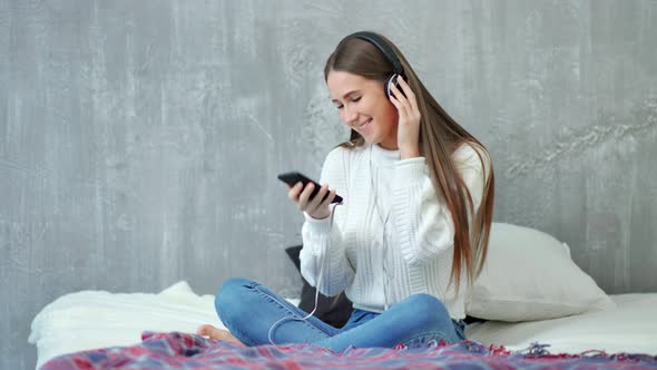Happy Teenage Woman Smiling Enjoying Listening Music Using Smartphone and Headphones Sitting on Bed