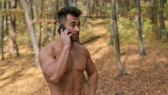 Strong Guy With Naked Torso Stands In Autumn Park And Speaks On Mobile Phone.