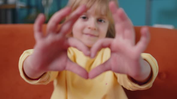 Smiling Child Girl Kid on Home Sofa Looking at Camera Makes Heart Gesture Demonstrates Love Sign