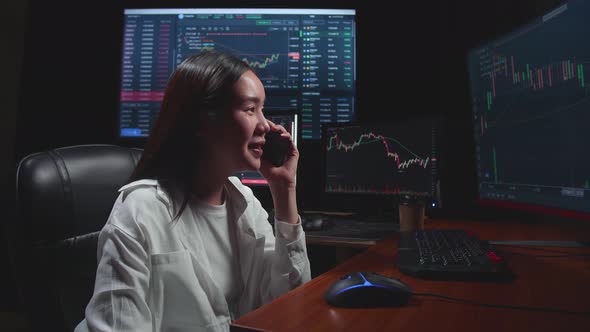 Young Female Stock Market Broker Talking On Mobile Phone With Analysing Graphs On Multiple Computer