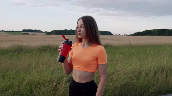 Young Girl Drinks From Sport Flask and Looks Thoughtfully at Camera on Nature