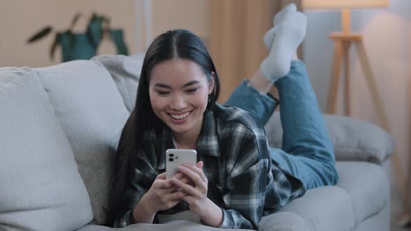 Asian Korean Girl Japanese Chinese Woman Lying Resting on Home Sofa Smiling Looking at Mobile Phone