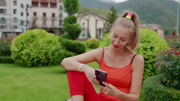 a Blonde in Red Clothes is Sitting with a Phone in Hands Against the Background of Greenery a Hotel