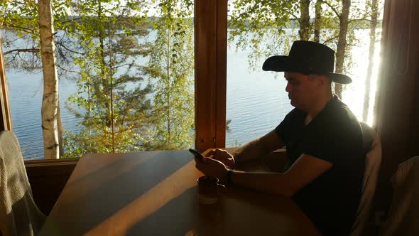 Man Using Smartphone Sitting at Table by Window of Cottage