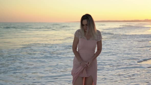 A Young Beautiful Blond Woman in a Pink Dress Walks Along the Beach at Sunset