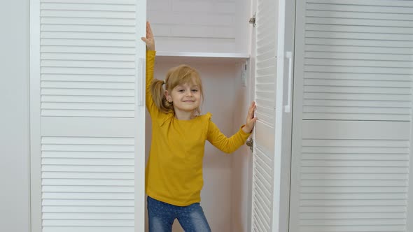 Little Blonde Child Girl Wearing Jeans, Orange Jacket Opening Wardrobe and Looking at Camera at Home