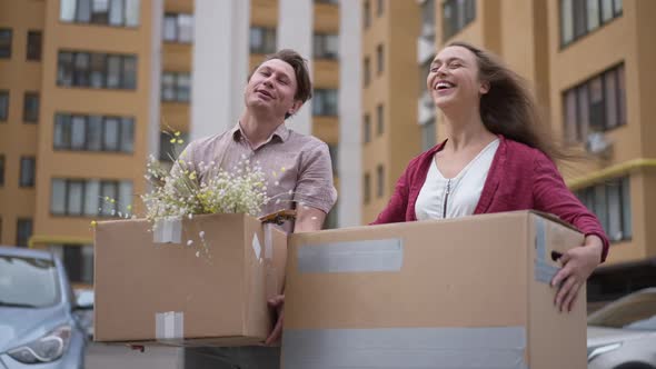 Excited Young Couple Standing with Boxes at Parking Lot Admiring House and Looking at Camera Smiling