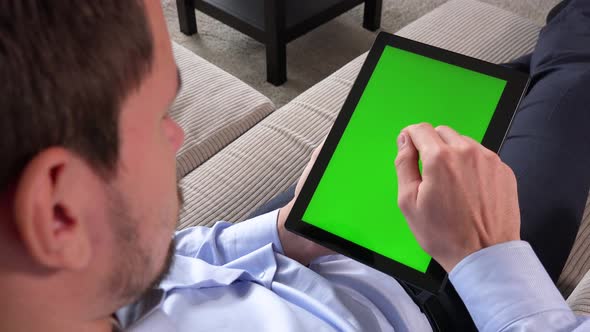 A Man Sits on a Couch and Works on a Tablet with a Green Screen in Vertical Position - He Taps