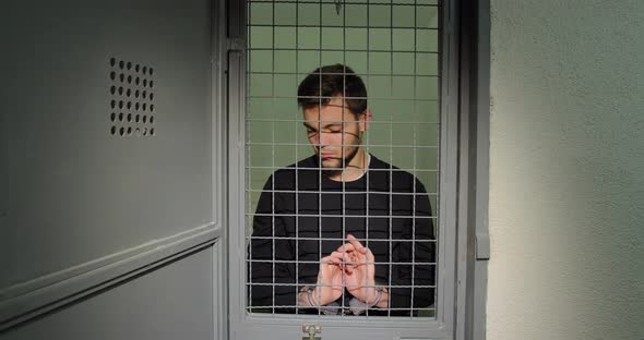 A Man In Handcuffs In A Prison Cell Stands Upset Holding On To A Lattice. Offender