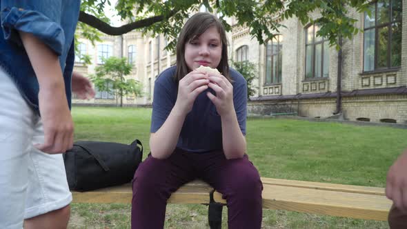 Relaxed Plump Boy Sitting on Bench with Sandwich As Unrecognizable Aggressive Classmates Knocking