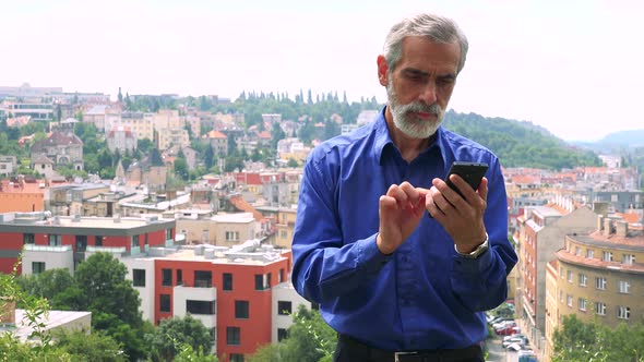 Old Senior Man Works (Typing) on Smartphone - City (Buildings) in Background