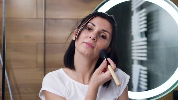 Young Brunette which Applying Face Powder with Brush and Looking at Camera