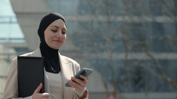 Muslim Woman Carrying Clipboard and Using Mobile Outdoors