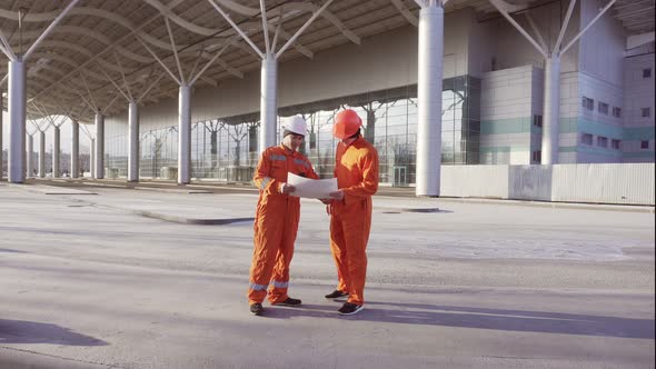 Two Construction Workers in Orange Uniform and Hardhats Meeting Each Other at the Bulding Object and