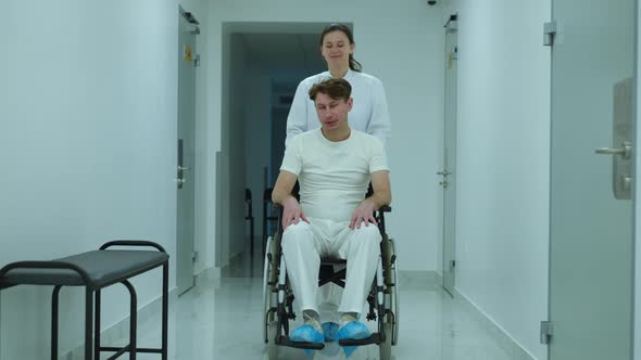 Wide Shot Portrait of Positive Doctor Talking to Patient Pushing Wheelchair in Hospital Hallway