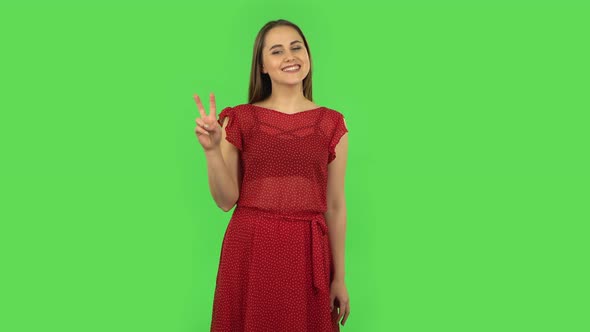 Tender Girl in Red Dress Is Showing Two Fingers Victory Gesture. Green Screen