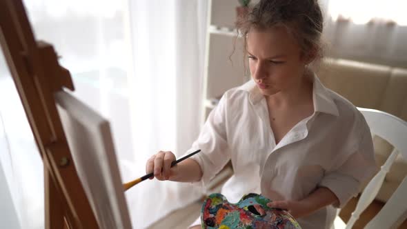 Pretty Girl in a White Shirt with Brush in Hand Sits Near an Easel and Draws on Canvas