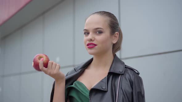 Portrait of a Cute Girl in an Emerald Evening Dress and a Leather Jacket Biting a Juicy Apple. Real