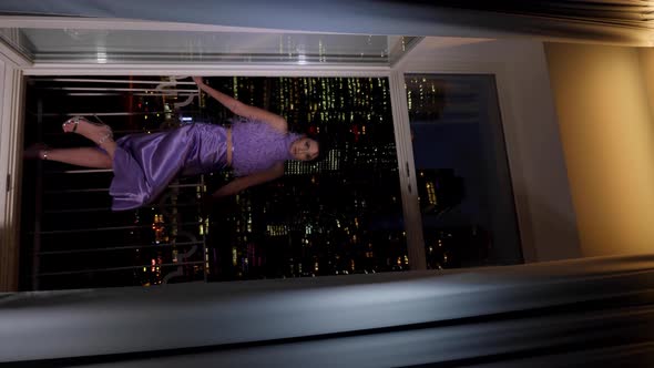 Rich Woman in a Purple Dress Standing By the Window and Looks at the Night City with Skyscrapers