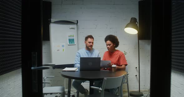 Two Young Colleagues Working in a Small Meeting Room with a Glass Wall and Door