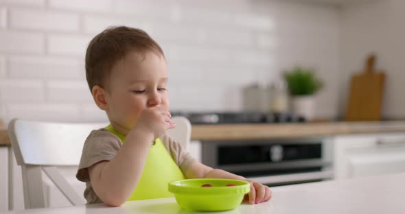 Cute Little Baby Boy Sits at a Table in the Kitchen and Eats Strawberries From Green Plate with His