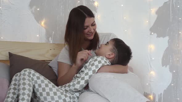 A Beautiful Mother Plays with Her Son Sitting on the Bed and Kisses Him Having a Good Mood