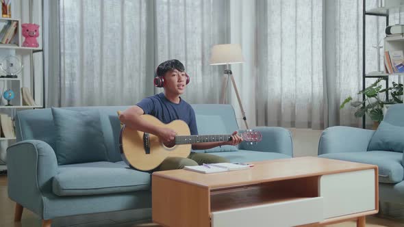 Asian Boy Composer With Notebook On Table Wearing Headphones Singing And Playing Guitar At Home