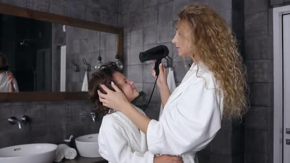 Blond Young Woman with Curly Hair in White Dressing Gown Drying Her Son's Hair in the Bathroom