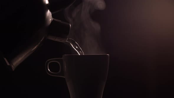 Hot boiled water is poured from a metal kettle into a white ceramic cup or mug on a black background