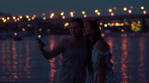 Two Girlfriends Do Selfie at Night By the River Against the Backdrop of Lights