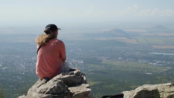A girl in an orange T-shirt sits on the edge of a cliff, looking out over the city and fields