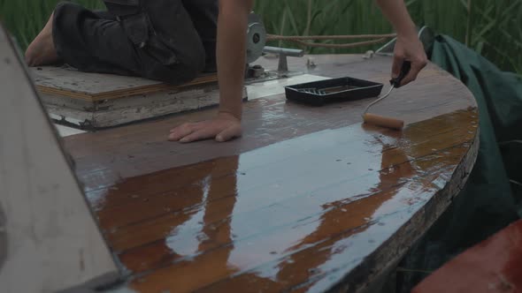 Young man sealing bow of wooden boat deck rolling on varnish.