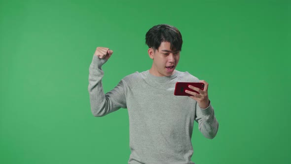 Asian Man Celebrating After Winning Game On Mobile Phone While Standing In The Green Screen Studio