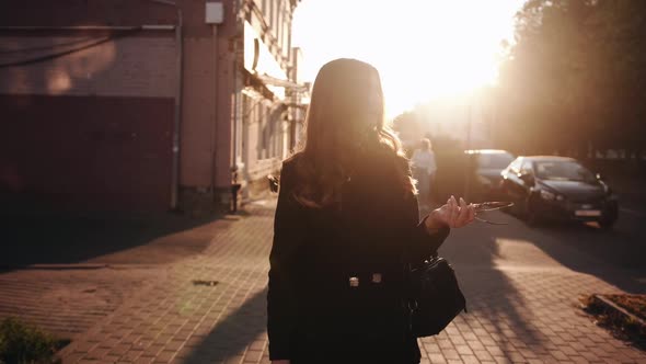 A Beautiful Young Girl in Stylish Black Clothes Goes Through the City at Sunset and Puts Sunglasses