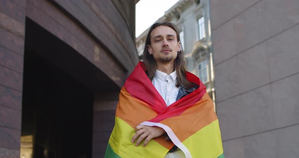 Portrait of Young Long Haired Man Covered Himself in Rainbow Flag While Looking To Camera