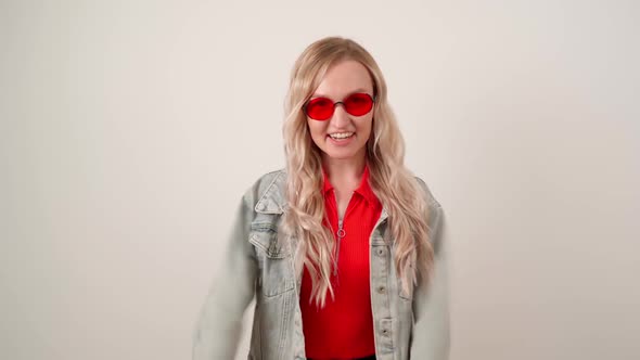 Happy Blonde in Red Glasses and a Denim Jacket is Dancing in Front of the Camera on a White