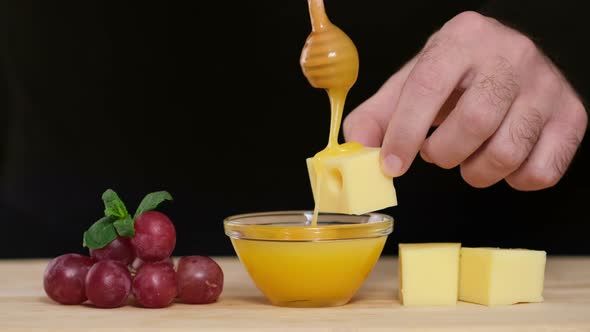 Man tasting cheese with honey and grapes. Dips wooden stick to glass bowl with liquid honey