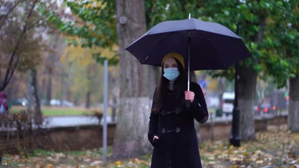 A Young Woman in a Protective Mask Walking in the Park Under Umbrella. Rainy Day, During Second Wave