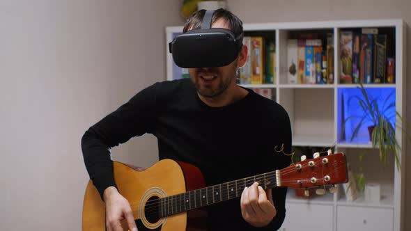 A man wearing virtual reality glasses sings while playing an acoustic guitar sitting in a room