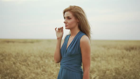 Handsome Young Woman in a Long Blue Dress Standing in Golden Wheat Field Trying the Wheat's Stem