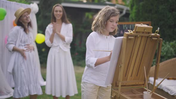 Concentrated Teenage Girl Painting on Easel Outdoors with Blurred Pregnant Mother Talking with