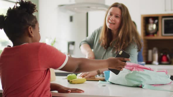 Happy caucasian woman and her african american daughter using smartphone in kitchen