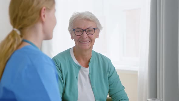 Careful nurse with ponytail talks to senior woman resident and gives glass of water