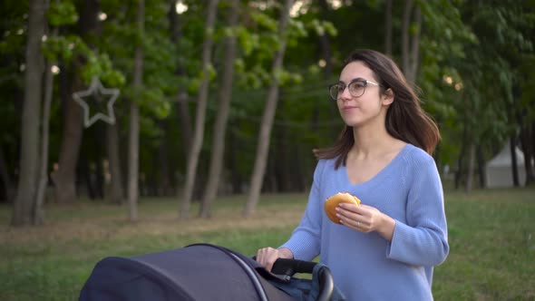 A Young Woman in Glasses Walks with a Stroller in the Park and Eats a Burger